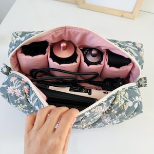May include: A floral-patterned cosmetic bag with a pink interior, open to reveal hair styling tools. The bag is gray with a white zipper and features a quilted design with pink and white floral accents. Inside are hairbrushes and a curling iron.