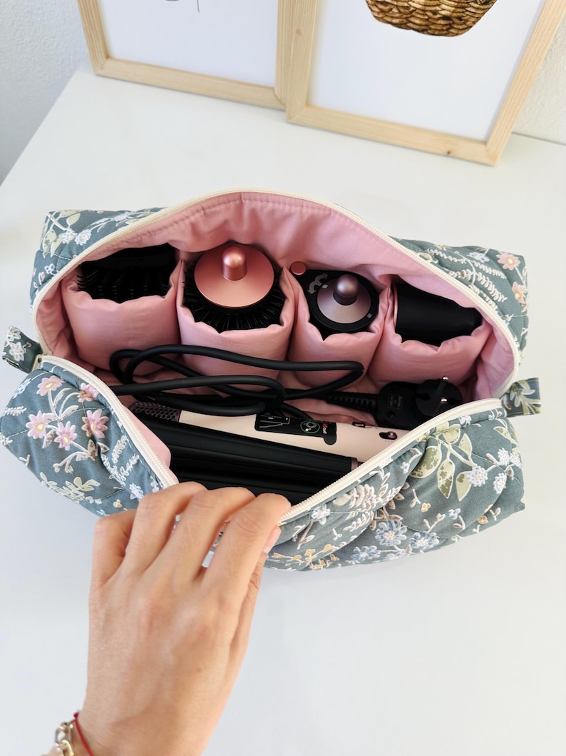 May include: A floral-patterned cosmetic bag with a pink interior, open to reveal hair styling tools. The bag is gray with a white zipper and features a quilted design with pink and white floral accents. Inside are hairbrushes and a curling iron.