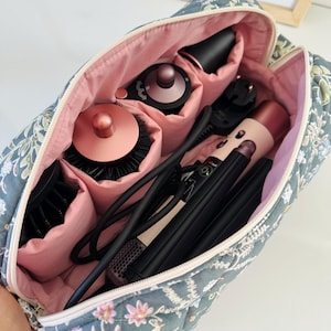 May include: A floral-patterned travel bag, open to reveal hair styling tools. The bag is quilted in a blue-grey fabric with pink floral accents and a pink interior. Inside are various black and rose gold hair styling tools, including a hair dryer and curling iron.