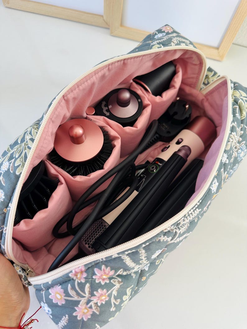 May include: A floral-patterned travel bag, open to reveal hair styling tools. The bag is quilted in a blue-grey fabric with pink floral accents and a pink interior. Inside are various black and rose gold hair styling tools, including a hair dryer and curling iron.