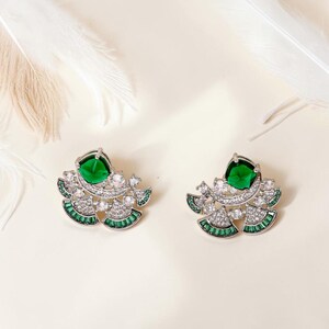 May include: A pair of silver-toned stud earrings featuring a central square-cut emerald green stone, surrounded by a fan-shaped arrangement of clear round crystals and small baguette-cut emerald green stones set in a metallic frame.