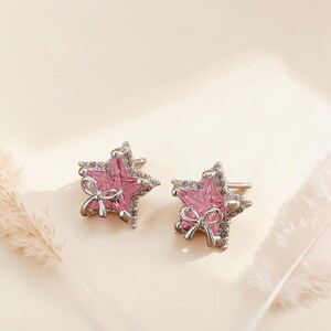 May include: A pair of silver stud earrings shaped like stars, featuring a central pink gemstone, a small silver bow accent at the bottom, and a border of tiny clear pavé stones set into the metal frame, creating a sparkling and detailed design.