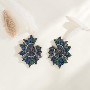 Könnte beinhalten: Ein Paar fächerförmige Ohrringe mit einem Spiraldesign, das mit kleinen blauen, grünen und klaren Kristallen auf einem dunklen Metallrahmen besetzt ist. Das geometrische, strahlenförmige Muster erzeugt ein markantes, geschichtetes Aussehen.