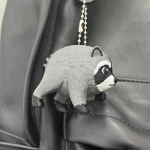 May include: A small, gray and black raccoon figurine keychain attached to a silver-colored ball chain. The raccoon has a rounded body, pointed ears, and a distinct black mask around its eyes. It is hanging from a dark gray backpack strap.