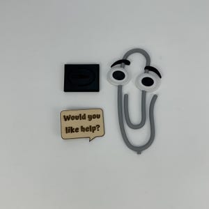 May include: A gray paperclip with cartoon eyes, a black square, and a wooden speech bubble that reads "Would you like help?" on a white background. Office supplies.