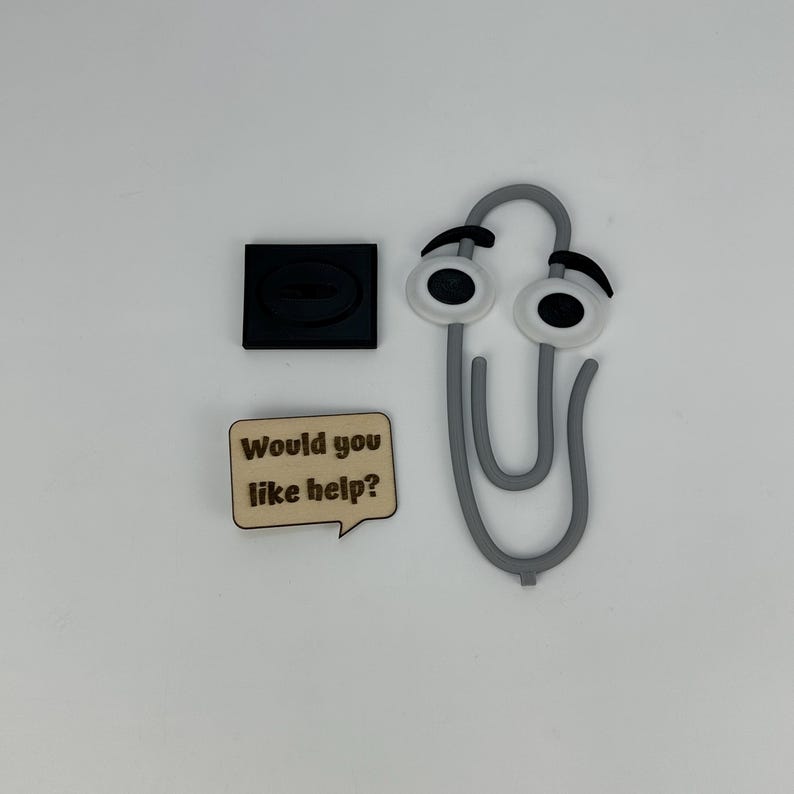 May include: A gray paperclip with cartoon eyes, a black square, and a wooden speech bubble that reads "Would you like help?" on a white background. Office supplies.