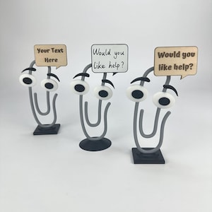 May include: Three whimsical paper clip desk organizers with googly eyes and speech bubbles. One bubble reads "Your Text Here," and two say "Would you like help?" The clips are gray, with black bases.
