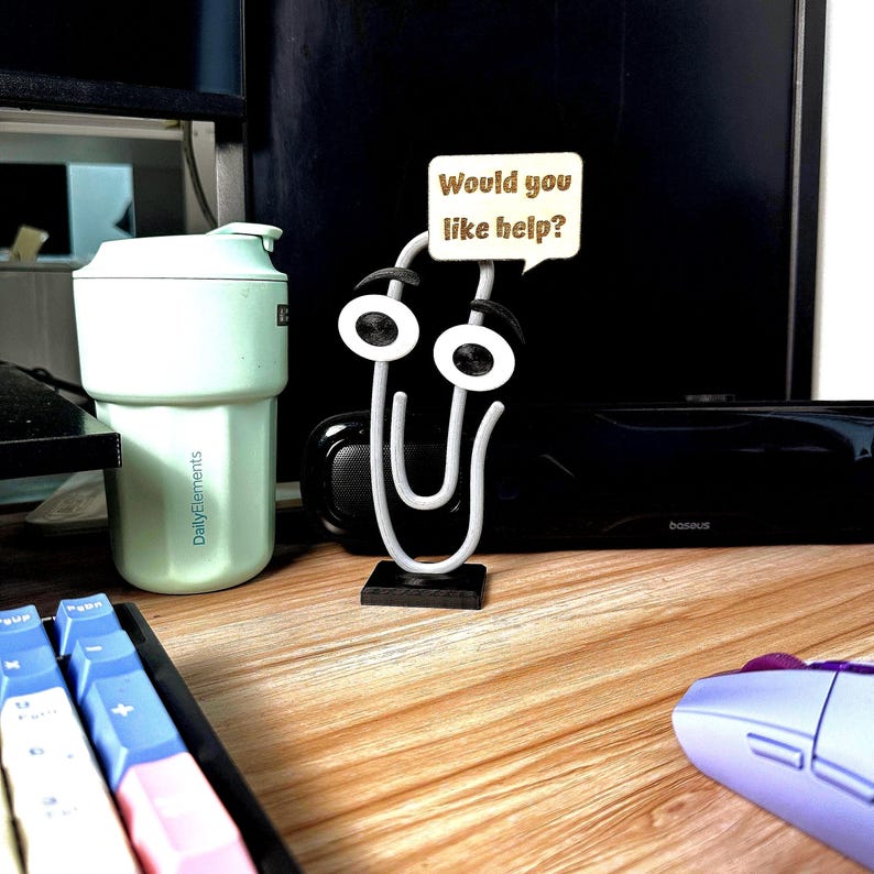 May include: A whimsical paperclip-shaped desk accessory with a speech bubble that reads "Would you like help?". The paperclip has white eyes and black accents. A mint green travel mug and a keyboard are also visible.