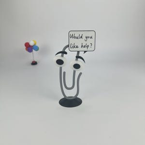May include: A whimsical, gray paperclip with googly eyes and a small sign that reads "Would you like help?" The paperclip is attached to a black base. In the background, a small decorative item with colorful spheres.