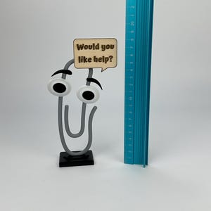May include: A whimsical paperclip-shaped desk accessory with googly eyes and a speech bubble that reads "Would you like help?". A blue ruler is next to the paperclip. The paperclip is approximately 3 inches tall.