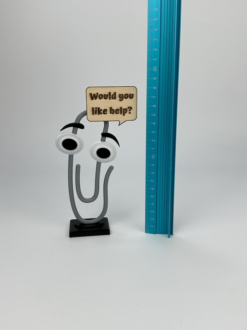 May include: A whimsical paperclip-shaped desk accessory with googly eyes and a speech bubble that reads "Would you like help?". A blue ruler is next to the paperclip. The paperclip is approximately 3 inches tall.