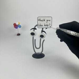 May include: A whimsical gray paperclip figure with googly eyes and a speech bubble that reads "Would you like help?" A hand in a black glove holds a whiteboard marker. A small colorful balloon decoration is in the background.