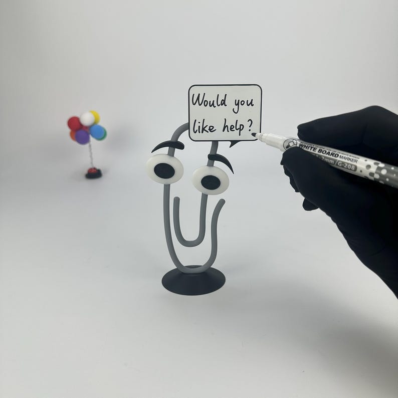 May include: A whimsical gray paperclip figure with googly eyes and a speech bubble that reads "Would you like help?" A hand in a black glove holds a whiteboard marker. A small colorful balloon decoration is in the background.