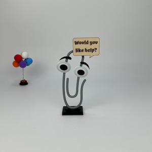 May include: A whimsical gray paperclip character with googly eyes and a speech bubble that reads "Would you like help?" The paperclip is mounted on a black base. A small colorful balloon arrangement is in the background.