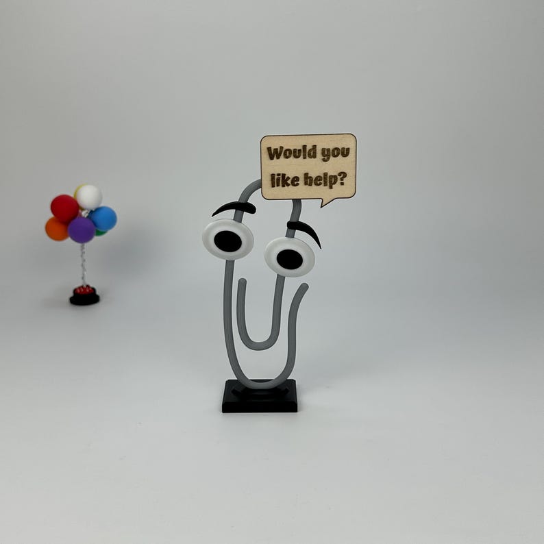 May include: A whimsical gray paperclip character with googly eyes and a speech bubble that reads "Would you like help?" The paperclip is mounted on a black base. A small colorful balloon arrangement is in the background.