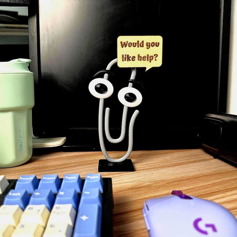 May include: A whimsical desk accessory featuring a large, silver paperclip with googly eyes and a speech bubble that reads "Would you like help?". The paperclip is mounted on a black base, suggesting a playful office or workspace theme.