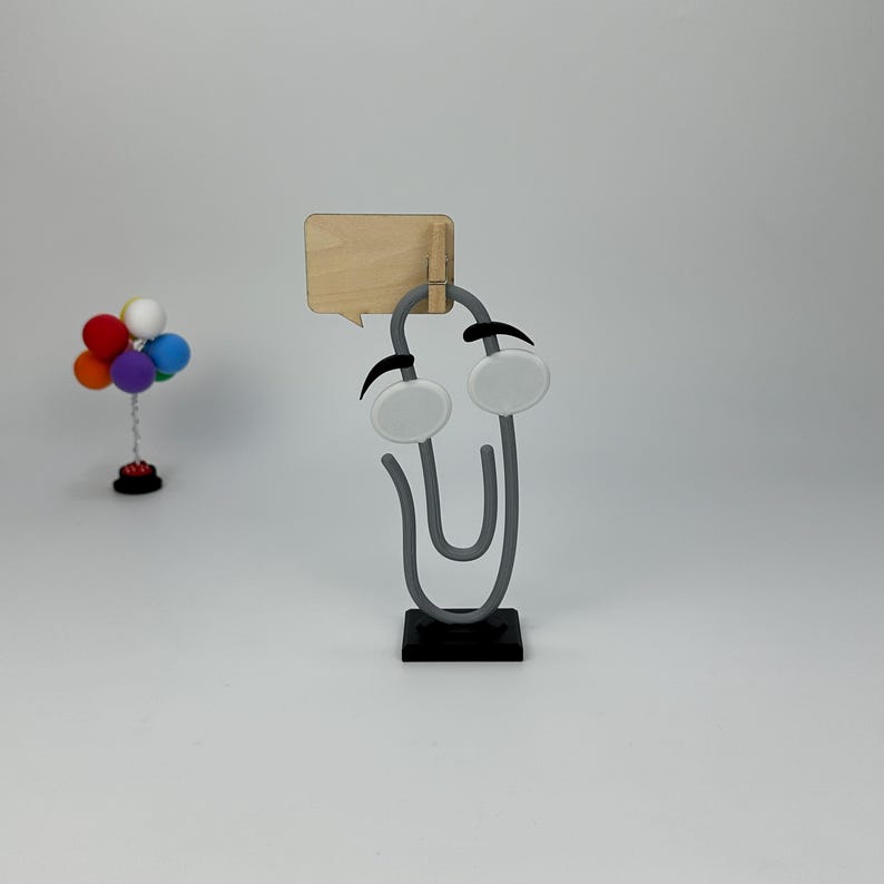 May include: A whimsical gray paperclip character with white eyes and a wooden speech bubble. A small colorful balloon arrangement is in the background. The paperclip is on a black base.
