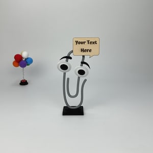 May include: A whimsical gray paperclip with googly eyes and a wooden speech bubble that reads "Your Text Here." A small colorful balloon arrangement is in the background. The paperclip is on a black base.