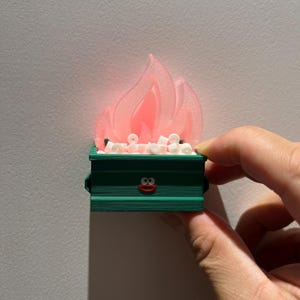 May include: A green miniature dumpster with a smiling face and a pink flame on top. White marshmallows are inside the dumpster.