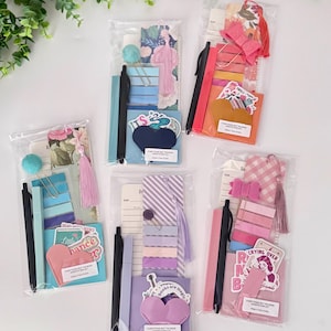 May include: Five clear cellophane bags filled with stationery items. Each bag contains a pen, sticky notes, stickers, and a bookmark. The bags are arranged on a white surface with green foliage in the background. The stickers have text that reads "Everything But The Book Annotation Kit".