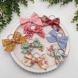 May include: A collection of fabric bow keychains in various patterns and colors, including pink gingham, floral prints in red, blue, and yellow, and a blue and white checkered design. The bows are arranged on a white, round tray.