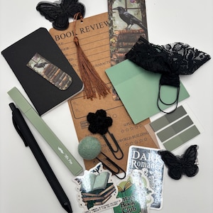 K&ouml;nnte beinhalten: Eine Auswahl an Schreibwaren in Schwarz-, Gr&uuml;n- und Braunt&ouml;nen, darunter ein Notizbuch, Aufkleber, Lesezeichen und ein Stift. Die Aufkleber zeigen die Texte "Fantasy Book Club" und "Dark Romance". Das Thema umfasst Schmetterlinge und Raben.