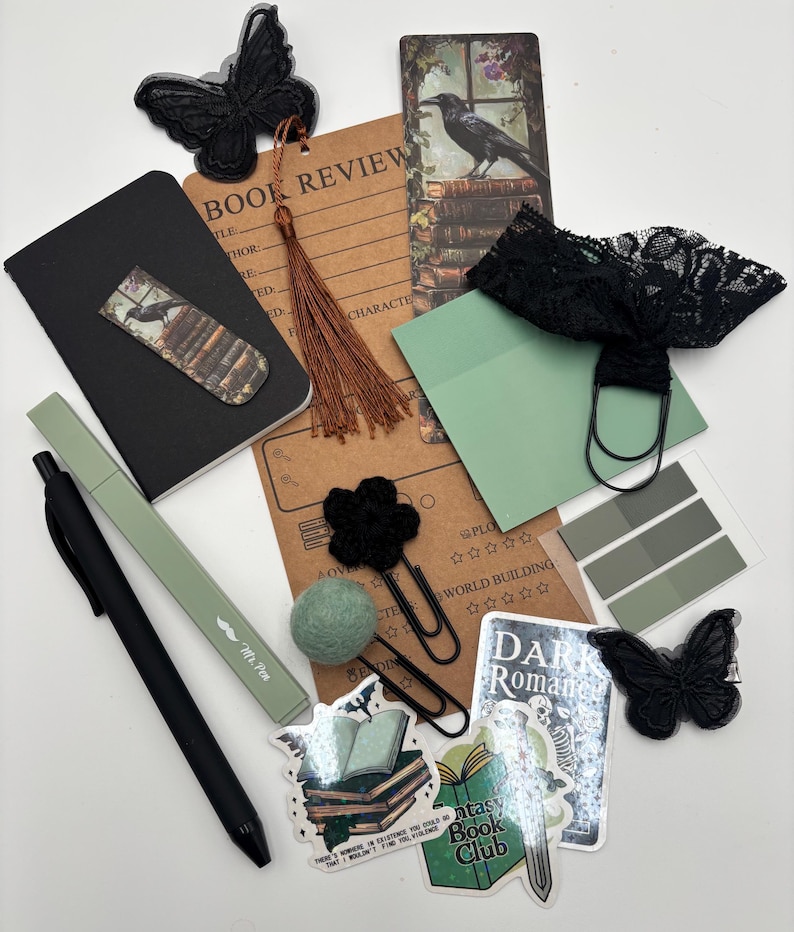 K&ouml;nnte beinhalten: Eine Auswahl an Schreibwaren in Schwarz-, Gr&uuml;n- und Braunt&ouml;nen, darunter ein Notizbuch, Aufkleber, Lesezeichen und ein Stift. Die Aufkleber zeigen die Texte "Fantasy Book Club" und "Dark Romance". Das Thema umfasst Schmetterlinge und Raben.