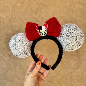 May include: Crocheted headband with gray ears and a red bow. The bow features a black and white character emblem. The headband is black. The ears are a speckled gray and white.