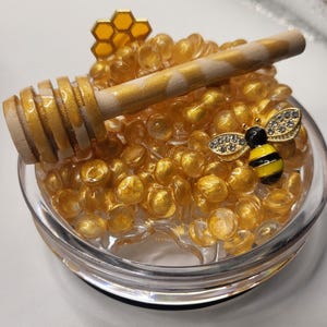 May include: A clear glass dish filled with golden, round beads. A wooden honey dipper with gold accents rests on top. A small, jeweled bee figurine and a honeycomb-shaped charm add decorative elements to this honey-themed display.