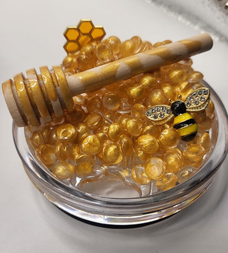 May include: A clear glass dish filled with golden, round beads. A wooden honey dipper with gold accents rests on top. A small, jeweled bee figurine and a honeycomb-shaped charm add decorative elements to this honey-themed display.