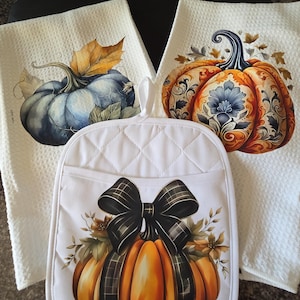 May include: White kitchen towels and a pot holder featuring pumpkin designs. The towels display watercolor pumpkins in blue and orange with autumn leaves. The pot holder has a pumpkin with a black plaid bow.