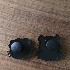May include: Two black enamel pins with cartoon cat designs. One pin is a cat head with a bow, the other a cat silhouette. Both pins have a black rubber clutch on the back for attachment.