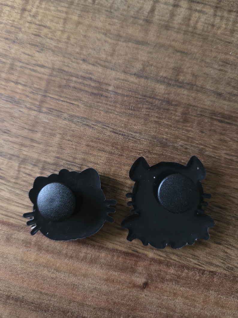 May include: Two black enamel pins with cartoon cat designs. One pin is a cat head with a bow, the other a cat silhouette. Both pins have a black rubber clutch on the back for attachment.