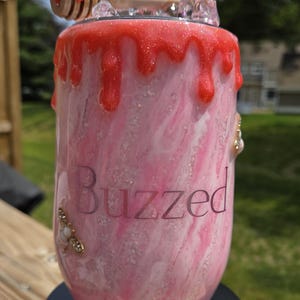 May include: A pink and white tumbler with a red, glittery, dripped design. The word "Buzzed" is printed on the side. The tumbler is decorated with a honey dipper, a honeycomb, and bee-themed embellishments.