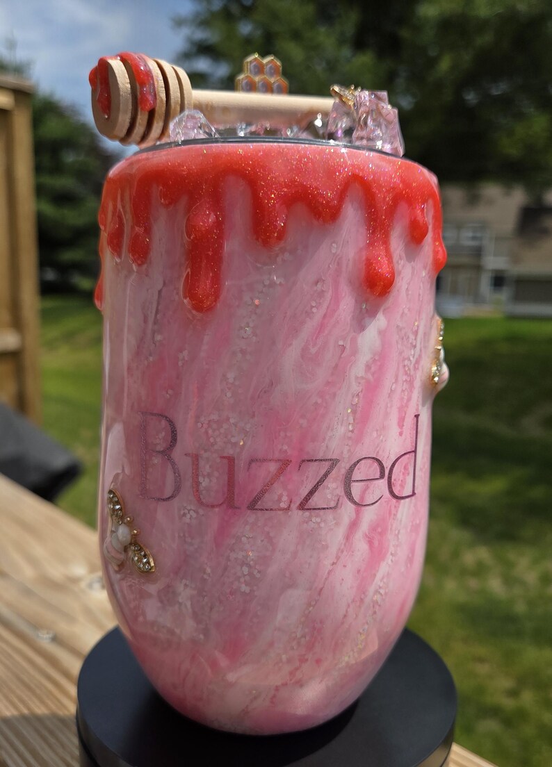 May include: A pink and white tumbler with a red, glittery, dripped design. The word "Buzzed" is printed on the side. The tumbler is decorated with a honey dipper, a honeycomb, and bee-themed embellishments.