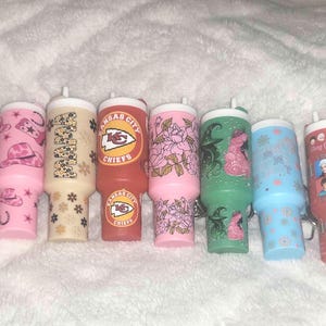 Könnte beinhalten: Eine Sammlung von 14 bunten Bechern mit verschiedenen Designs, darunter Comicfiguren, Sportlogos und Blumenmuster. Die Becher sind alle ungefähr gleich groß und geformt und in einer Reihe auf einer weißen Oberfläche angeordnet.