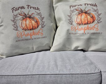 May include: Two square throw pillows with a fall theme. Each pillow features a watercolor-style orange pumpkin with a bow, surrounded by leaves. Text reads "Farm Fresh Pumpkins ALWAYS FRESH & PICKED DAILY."
