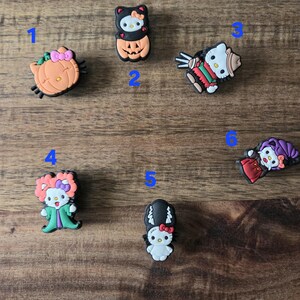 May include: A selection of six Halloween-themed shoe charms featuring Hello Kitty. The charms show Hello Kitty in various costumes, including a pumpkin, a witch, and a mummy. The charms are in various colours, including orange, black, and red.