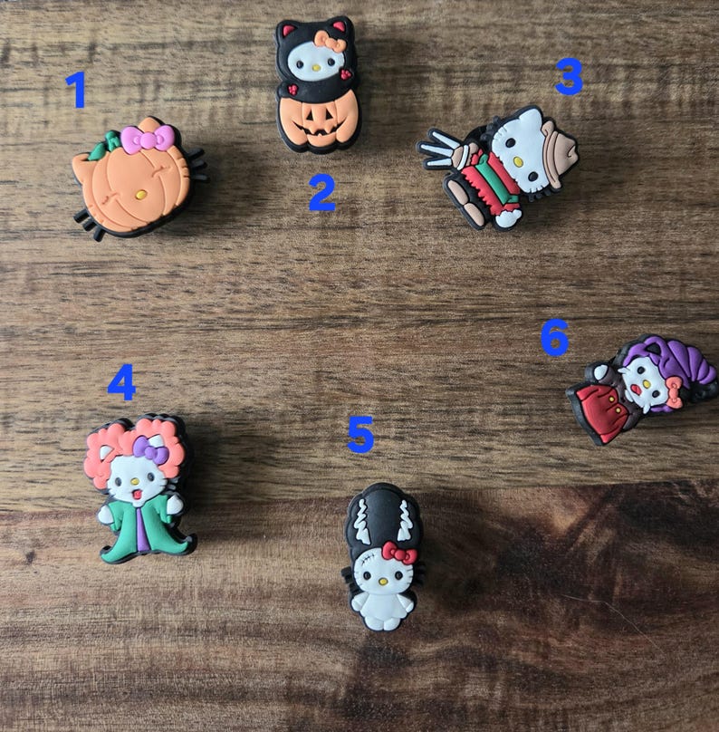 May include: A selection of six Halloween-themed shoe charms featuring Hello Kitty. The charms show Hello Kitty in various costumes, including a pumpkin, a witch, and a mummy. The charms are in various colours, including orange, black, and red.
