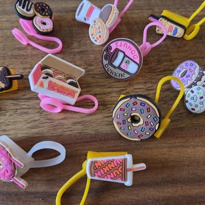 Op de afbeelding: Een verzameling kleurrijke bedels met voedselthema, waaronder donuts, koffiebekers en een doos donuts. De bedels hebben roze, bruine, gele en witte accenten. Elke bedel heeft een lus voor bevestiging.