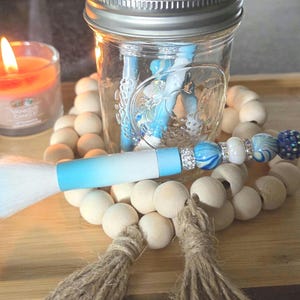 May include: A decorative makeup brush with a light blue handle and white bristles, resting on a wooden tray with a beaded garland and a lit candle. A glass jar with blue decorative items is in the background.