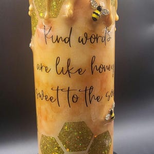 May include: A cylindrical tumbler with a honey-themed design. The tumbler features a golden, dripped honey effect, glittery honeycomb patterns, and decorative bee embellishments. The text "Kind words are like honey sweet to the soul" is printed on the tumbler.