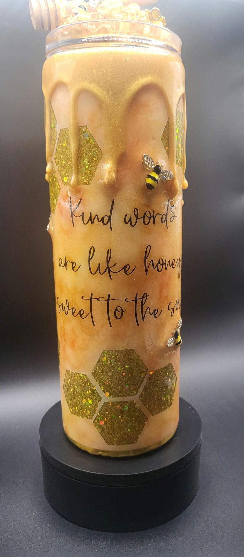 May include: A cylindrical tumbler with a honey-themed design. The tumbler features a golden, dripped honey effect, glittery honeycomb patterns, and decorative bee embellishments. The text "Kind words are like honey sweet to the soul" is printed on the tumbler.