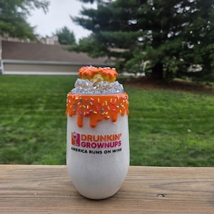 May include: A white tumbler with a glitter finish featuring the text "Drunkin' Grownups" and "America Runs on Wine" in orange and pink. The rim has orange icing drip with colorful sprinkles, and a miniature donut sits on top of clear ice cubes.