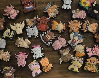 May include: A selection of colourful Highland cow shoe charms in various designs. Some charms feature the cows with books, wings, and flowers. Others have hearts and bows. The charms are in shades of brown, pink, white, and yellow.