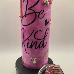 May include: A pink tumbler with the words "Be Kind" in black script. The tumbler has a dripping pink design at the top, with gold bee embellishments. A small glass container with pink beads and a honey dipper is next to the tumbler.
