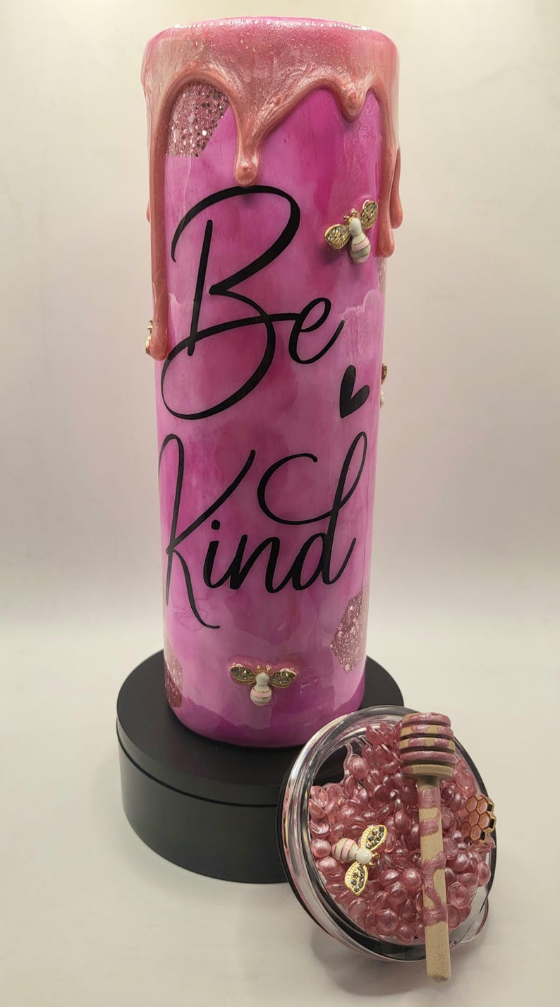 May include: A pink tumbler with the words "Be Kind" in black script. The tumbler has a dripping pink design at the top, with gold bee embellishments. A small glass container with pink beads and a honey dipper is next to the tumbler.