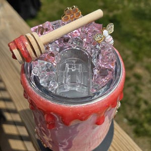 May include: A custom-made tumbler with a honey-themed design. The tumbler features a pink and white marble effect, topped with pink crystals, a wooden honey dipper, and bee-themed embellishments. The rim is decorated with a red, glittery drip effect.
