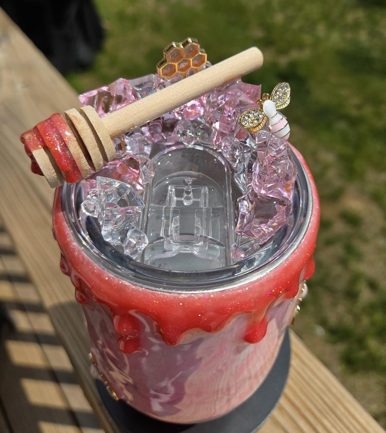 May include: A custom-made tumbler with a honey-themed design. The tumbler features a pink and white marble effect, topped with pink crystals, a wooden honey dipper, and bee-themed embellishments. The rim is decorated with a red, glittery drip effect.