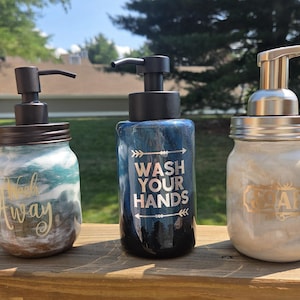 May include: Three glass soap dispensers with pump mechanisms. The left jar has a teal and white swirl pattern with the text Wash Away. The center jar is dark blue with the text WASH YOUR HANDS. The right jar is white with the text SOAP.